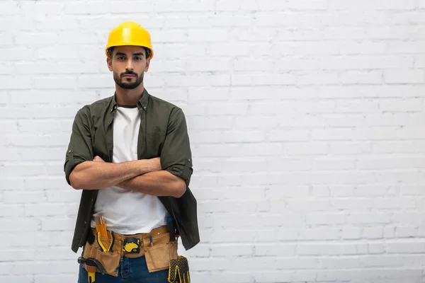 Arabian handyman in helmet and tool belt looking at camera at home — Stockfoto