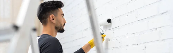 Side view of muslim man in glove coloring wall with roller near blurred ladder at home, banner — Stock Photo