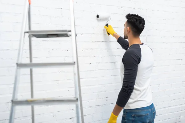 Arabian man in gloves coloring wall near ladder at home — Photo de stock