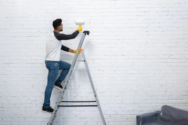 Arabian man in gloves coloring wall while standing on ladder at home — Foto stock