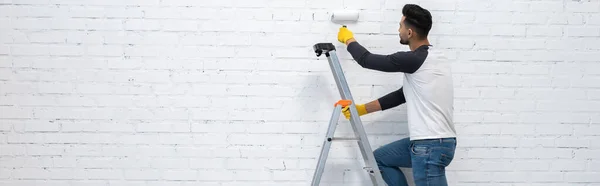 Muslim man in gloves coloring brick wall while standing on ladder at home, banner — Photo de stock
