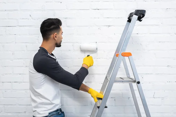 Young arabian man painting white brick wall and holding ladder at home — Fotografia de Stock