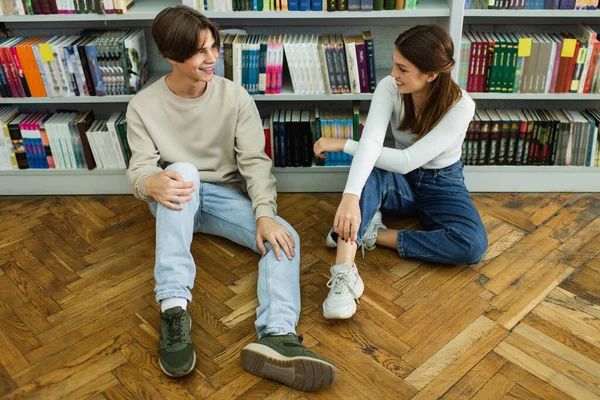 Cheerful teenagers talking and sitting on floor near bookshelves in library — Stock Photo