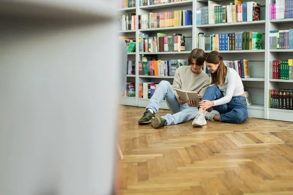 Couple of teenage students reading book in library while sitting on floor on blurred foreground — Stock Photo