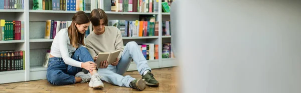 Full length of smiling teenage students reading book on floor on blurred foreground, banner — Stock Photo