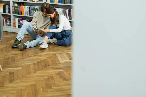 Happy teenage couple sitting on floor and reading book near racks in library on blurred foreground — Stock Photo