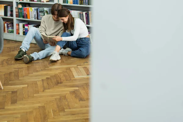 Счастливая пара подростков, сидящих на полу и читающих книги возле стойки в библиотеке на размытом переднем плане — стоковое фото