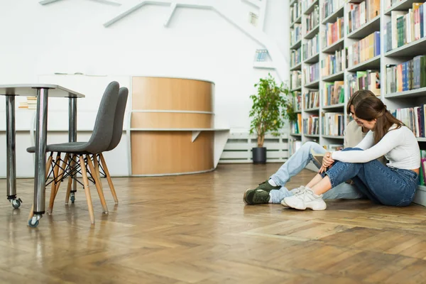 Подростковые друзья сидят на полу возле книжных полок и читают книги в библиотеке — стоковое фото