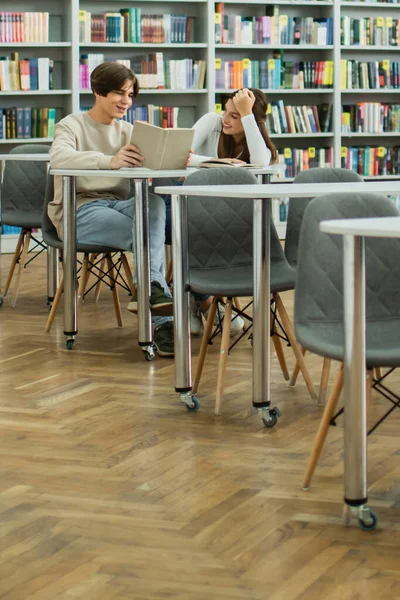 Студент читає книгу до усміхненої дівчини-підлітка, сидячи в бібліотеці — стокове фото