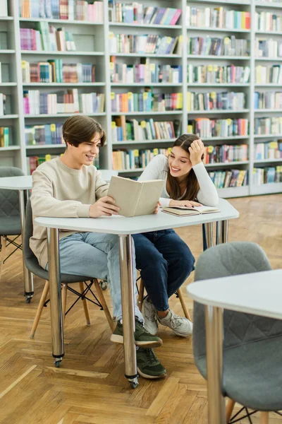 Teenage guy reading book near smiling friend in library — Photo de stock