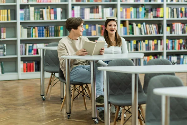 Девочка-подросток с книгой на голове рядом с улыбающейся подругой в читальном зале библиотеки — стоковое фото