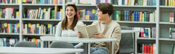 Cheerful teenage girl sitting with book on head near laughing friend in reading room, banner - foto de stock