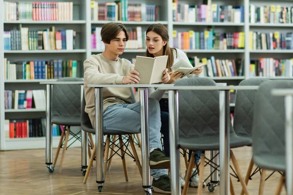 Student reading book near curious teenage girl in library - foto de stock