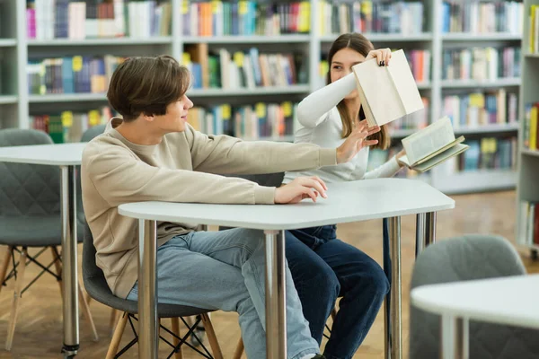 Smiling guy sitting near teen girl holding books in library reading room — Foto stock