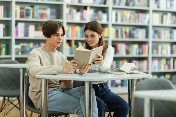 Улыбающаяся девочка-подросток показывает книгу другу, сидя в библиотеке — стоковое фото