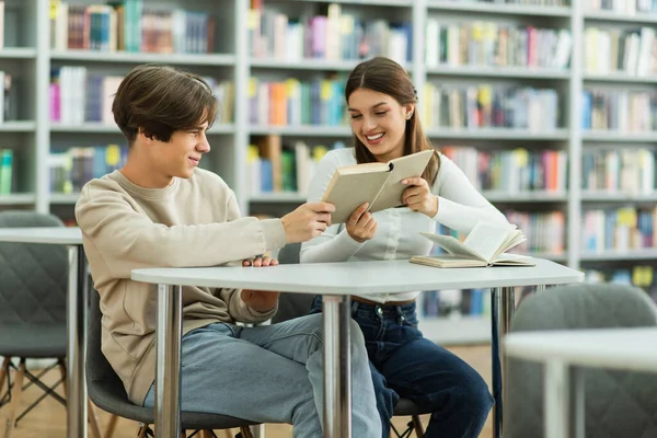 Cheerful teen girl showing book to smiling friend in library reading room — Stockfoto