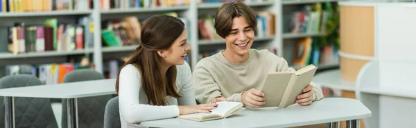 Cheerful student reading book near smiling teenage girl in library, banner — Photo de stock