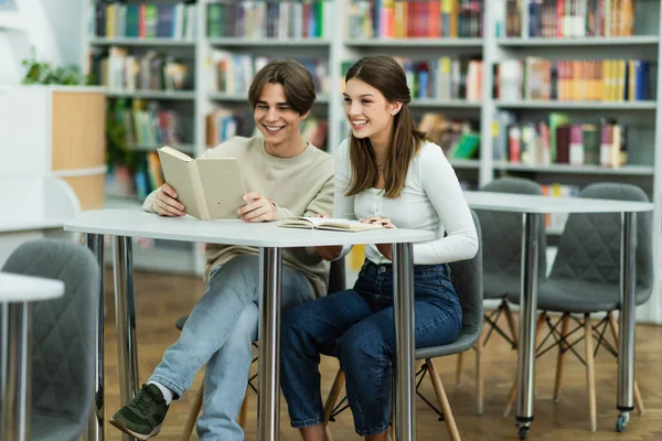 Smiling student reading book near cheerful teenage girl in library — Stock Photo
