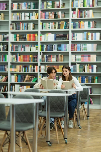 Teenage students reading books near bookshelves in library - foto de stock