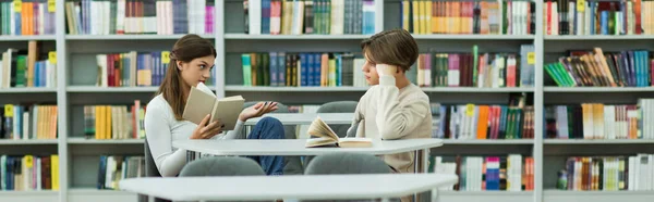 Teen girl holding book while talking to guy in library, banner — Photo de stock