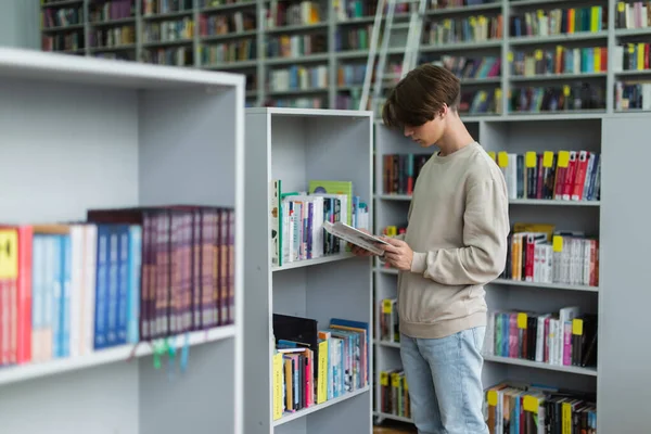 Тинейджер читает книги возле стойки в библиотеке — стоковое фото