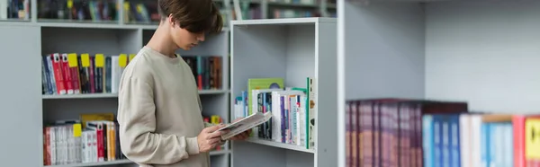 Teenager reading book while standing near racks in library, banner - foto de stock