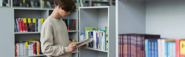 Подросток читает книгу, стоя возле стойки в библиотеке, баннер — стоковое фото