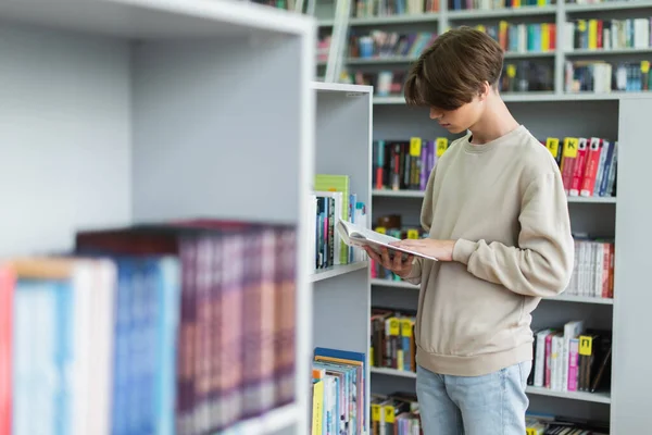 Студентка-підліток читає книгу біля книжкових полиць у бібліотеці на розмитому фоні — стокове фото