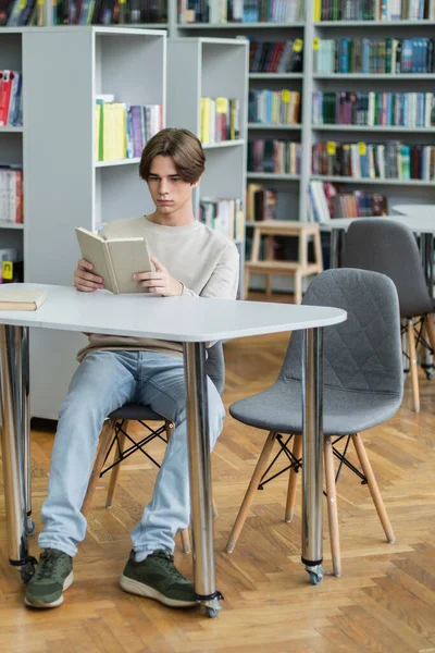 Full length of teenage student sitting at desk in library and reading book — Stock Photo