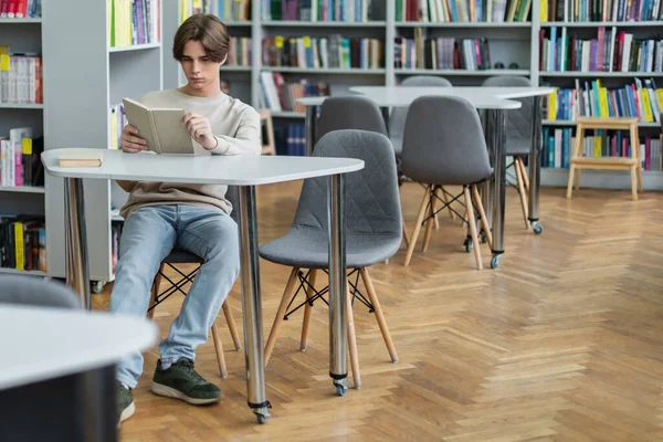 Teenage guy sitting with book in reading room of library - foto de stock