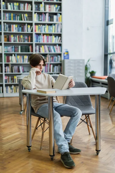 Full length of teenager sitting in library and reading book — Stock Photo