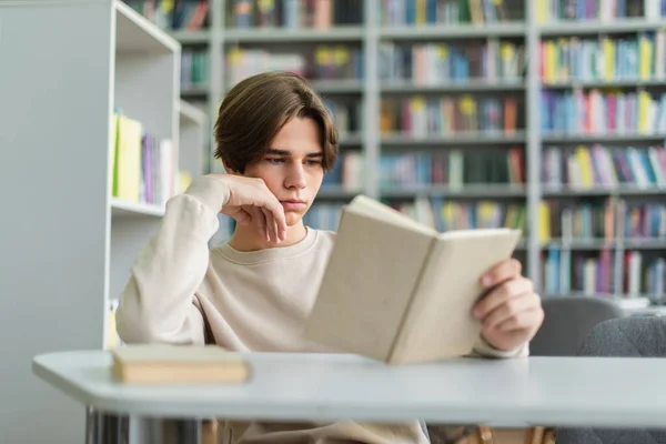 Teenage student reading book in library on blurred foreground - foto de stock