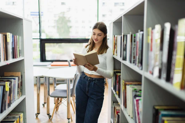 Teenage girl standing near shelves in library and reading book - foto de stock