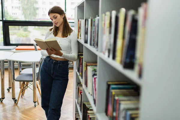 Teenage student reading book while standing near racks on blurred foreground - foto de stock