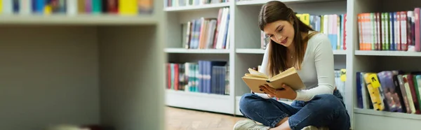 Студент-подросток, сидящий на полу в библиотеке и читающий книгу на размытом переднем плане, баннер — стоковое фото