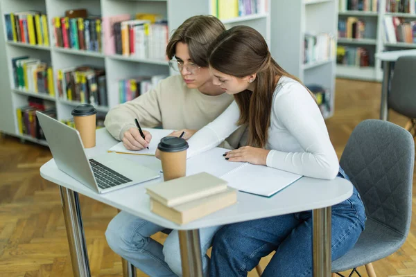 Teenage students writing near laptop and books in library - foto de stock