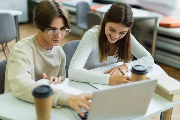 Smiling teenage girl writing near friend using laptop in library reading room — Stockfoto