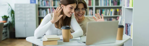 Smiling teenage students waving hands during video chat on laptop in library, banner - foto de stock