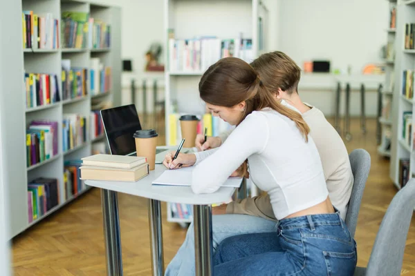 Side view of teenage girl writing near laptop with blank screen and friend in library — Foto stock