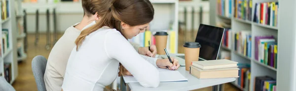 Side view of teen girl writing in notebook near friend and laptop with blank screen in library, banner — Stock Photo