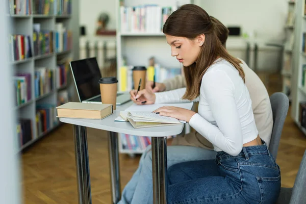 Teenage girl reading book near friend and laptop with blank screen in library — Stock Photo