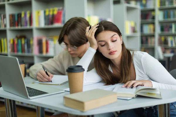 Teenage girl reading book near friend writing in notebook and laptop in library — Stock Photo