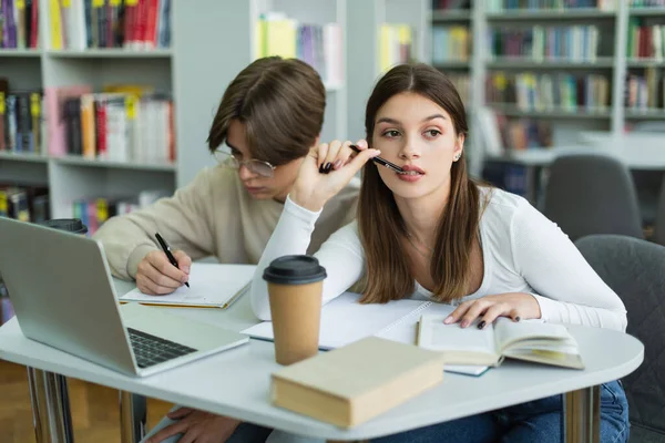 Thoughtful teen girl looking away near laptop and friend writing in notebook - foto de stock