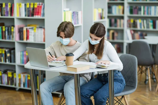 Подростки в медицинских масках учатся возле ноутбука и книжных полок в библиотеке — стоковое фото