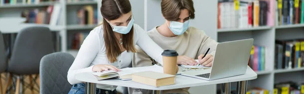 Guy in medical mask writing near teenage girl and laptop in library, banner - foto de stock