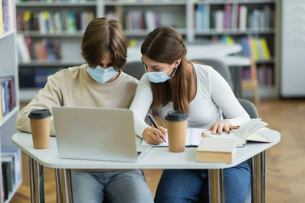 Teenage students in medical masks studying near laptop in library reading room — Stock Photo