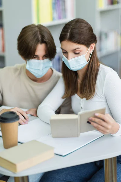 Teenage girl in medical mask reading book near paper cup and blurred friend — Stock Photo