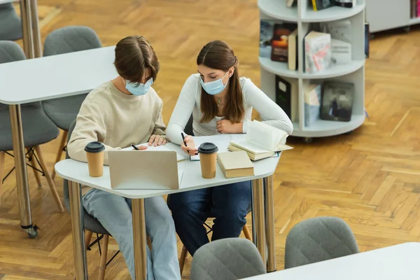 High angle view of teenage students in medical masks writing near laptop and paper cups in library - foto de stock