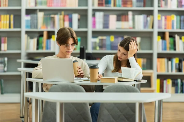 Teenage students with coffee to go sitting in library reading room near laptop - foto de stock