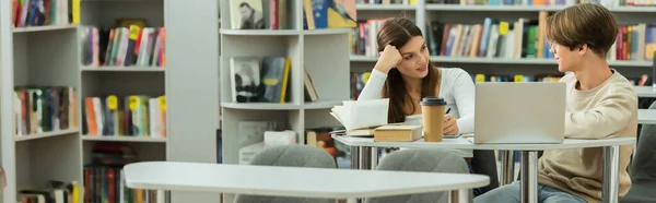 Усміхнена дівчина-підліток розмовляє з другом біля ноутбука і книг в бібліотеці, банер — стокове фото